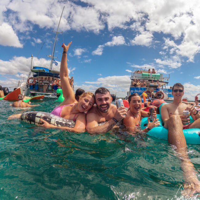 A group of smiling people in swimsuits enjoy drinks while floating in the ocean near boats under a partly cloudy sky, celebrating and raising their arms in a festive atmosphere.