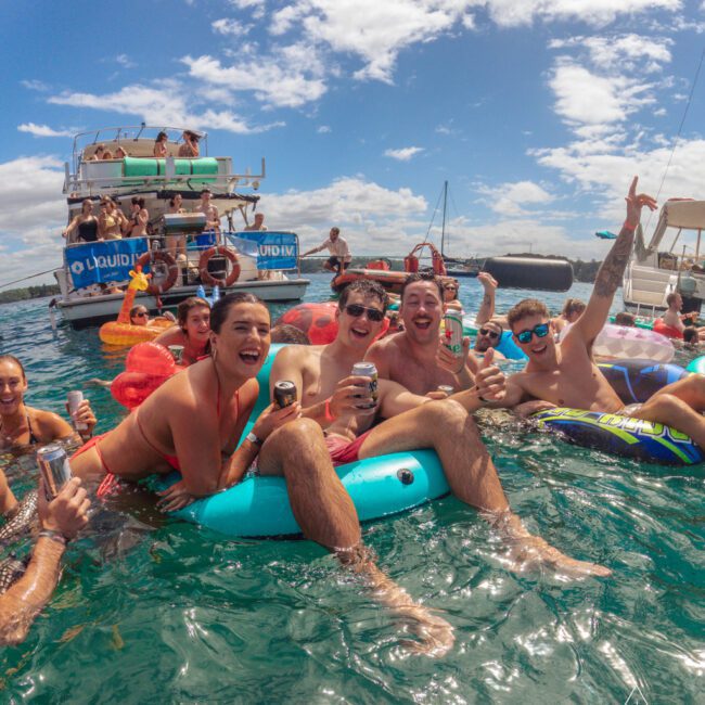 A group of young adults in swimsuits laugh and pose with drinks while floating on pool inflatables in the ocean, with boats and other partygoers in the background under a partly cloudy sky.