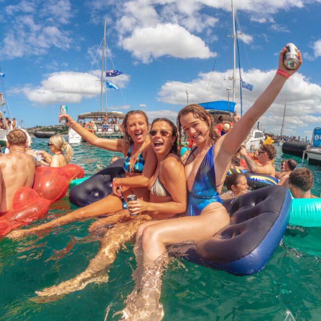 Three women in swimsuits smile and raise drinks while sitting on inflatable loungers in clear blue water. Others swim and relax nearby, with boats and a bright sky in the background. The atmosphere is lively and festive.