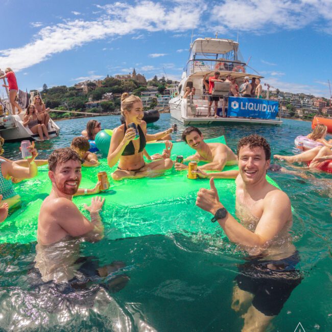 A group of young adults in swimsuits enjoy drinks while relaxing on a floating mat in the water near yachts on a sunny day; one man gives a thumbs-up to the camera.