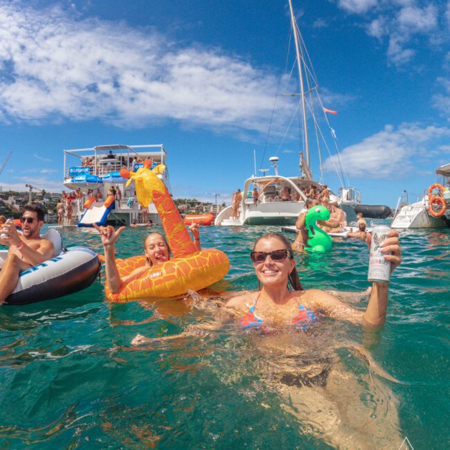 People enjoying a party in the ocean near boats under a sunny sky; a woman in sunglasses holds a drink, others float on inflatable pool toys shaped like animals.