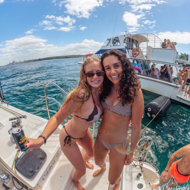 Two women in swimsuits smiling on a boat, with one holding a drink. Other people are relaxing on nearby boats in the water under a partly cloudy sky. The scene is lively and summery.