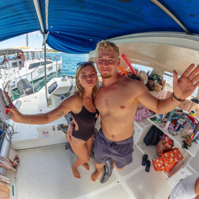 A smiling couple in swimwear pose for a selfie on a boat under a blue canopy, with various snacks and drinks visible around them. The sea and other boats are in the background.