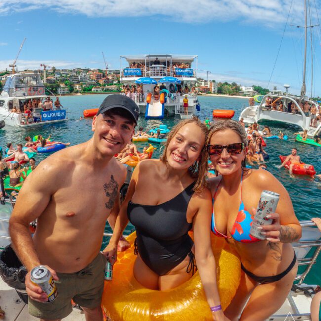 Three people in swimwear smile at the camera on a boat, holding drinks. Behind them, many others enjoy a lively boat party on the water, with floats, boats, and a sunny sky overhead.