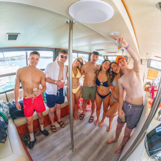 A group of seven young adults in swimsuits smile and pose together on a boat, holding drinks and enjoying a sunny day on the water. The lake and trees are visible through the windows behind them.
