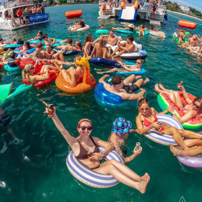 A group of young people relax on colorful inflatable floats in the ocean near boats on a sunny day, holding drinks and smiling. The scene is lively with others swimming and enjoying the water.