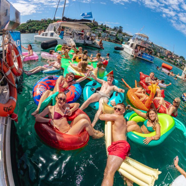 A group of people in swimsuits lounge on colorful inflatables in the water near anchored boats, smiling and enjoying a sunny day. The background shows more boats and a scenic shoreline under a partly cloudy sky.