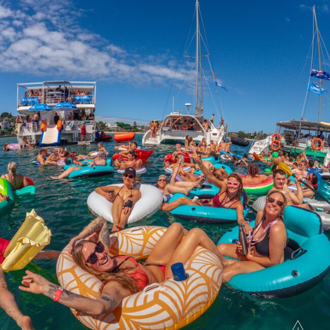 A group of people relax on colorful inflatable floats in the water, smiling and enjoying a sunny day. Boats are anchored in the background, and the sky is clear with scattered clouds.