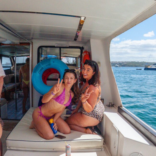 Two women in swimsuits smile and pose, making peace signs on a boat. Sunlight streams in, with snacks and drinks on a nearby table. The background shows clear blue water, boats, and a distant shoreline.