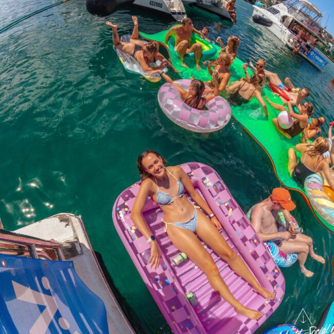 A woman in a bikini smiles while lounging on a pink float in clear water, surrounded by others relaxing on colorful inflatables near several docked boats under a sunny sky.
