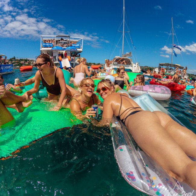 A group of people relax on colorful floaties in the water at a lively boat party. They’re smiling, laughing, and holding drinks, with boats and a clear blue sky in the background.