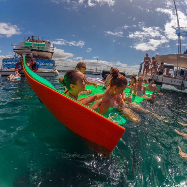 People are enjoying a party in the water near boats. They are using a long, orange floating device as a drink luge and are surrounded by bright green pool floats under a sunny sky with scattered clouds.