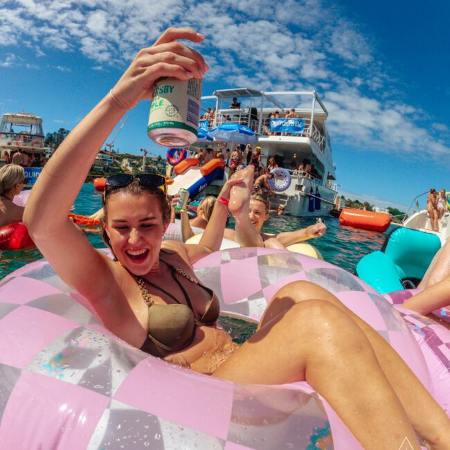 A woman in a bikini smiles and raises a can while lounging on a pink inflatable float in the water, surrounded by people enjoying a sunny boat party with yachts and blue skies in the background.