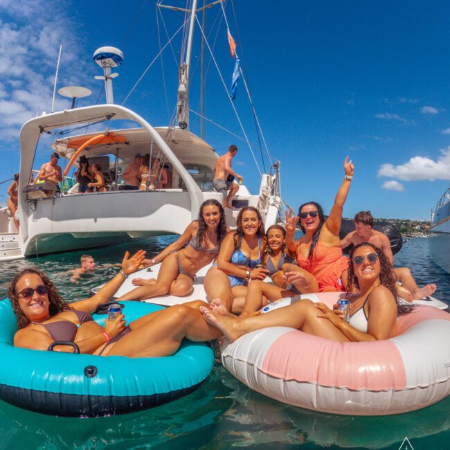 A group of smiling women in swimsuits relax on colorful pool floats in the ocean near a yacht, with other people enjoying the sunny day on the boat and in the water. The sky is clear and blue.