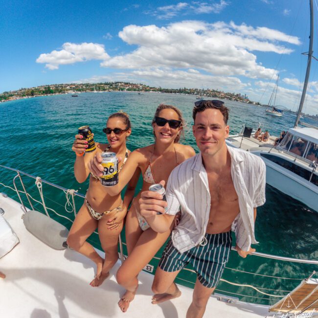 Three people in swimwear on a boat smile and hold up cans, with blue water, a sailboat, and a coastline with houses in the background under a sunny, partly cloudy sky.