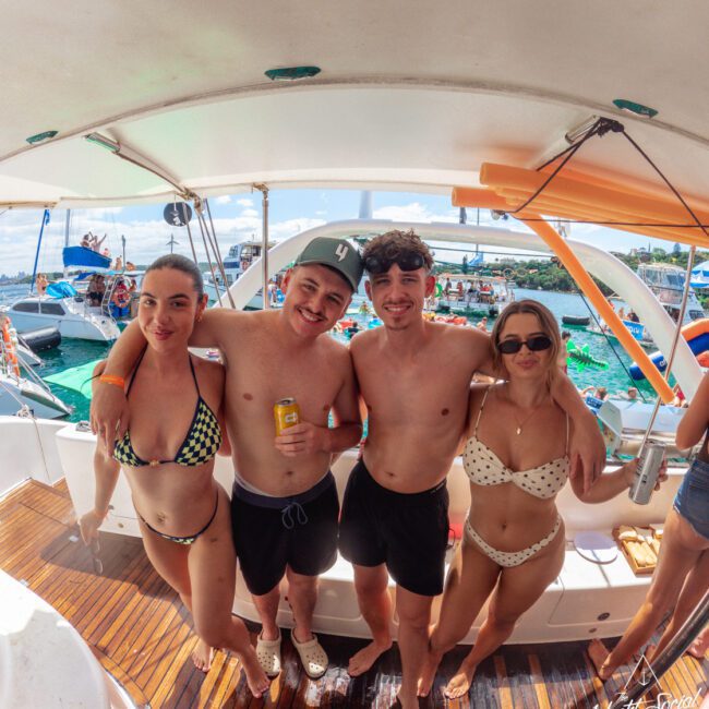Four young adults in swimsuits stand arm-in-arm, smiling on a boat with drinks in hand. Other boats and people are visible on the water in the background, suggesting a lively party atmosphere.