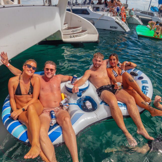 Four people in swimsuits relax on an inflatable raft in the water, smiling and holding drinks. Several boats and other people are visible in the background on a sunny day.