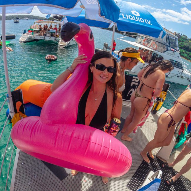 A smiling woman in sunglasses holds a drink and stands inside a pink flamingo float on a dock, surrounded by people in swimsuits. Boats and swimmers are in the turquoise water under a sunny sky.