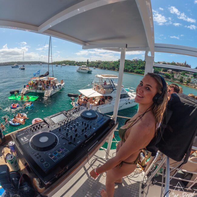 A woman in a bikini stands next to DJ equipment on a yacht, smiling at the camera. People are partying on boats and floating platforms on a sunny day with blue sky and water in the background.
