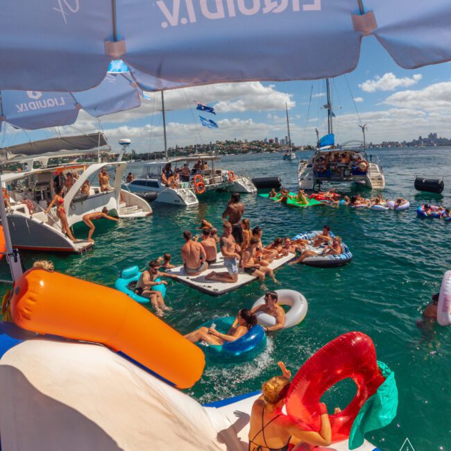 A group of people enjoy a lively yacht party in the ocean, lounging on colorful inflatables and swimming near several anchored boats under a sunny, blue sky. Large umbrellas provide shade on the boats.
