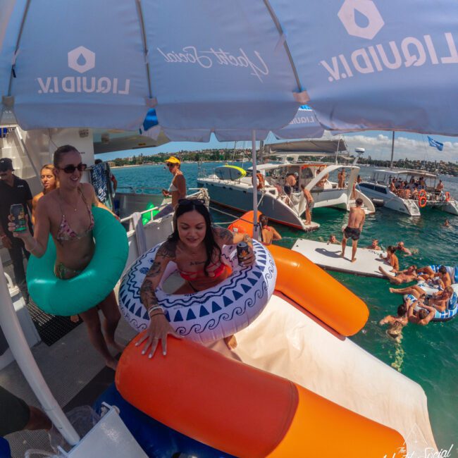 People enjoy a boat party on the water with inflatable tubes and a slide, while others swim nearby. Blue umbrellas provide shade and several boats are anchored close together under sunny skies.