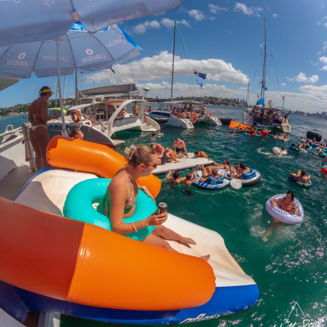 A woman relaxes on an inflatable pool float on a yacht, holding a drink. Nearby, people swim and float on tubes in turquoise water, with several boats anchored around under a sunny sky.