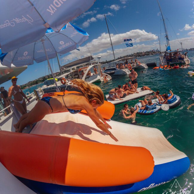 A woman in a blue bikini dives off a large inflatable slide on a yacht into the ocean, while people relax on inflatables and other boats nearby under sunny skies.