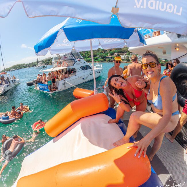 Two smiling women in swimsuits pose on an inflatable water slide on a yacht, with other people relaxing on nearby boats and in the water. The scene is sunny and lively, with umbrellas and clear blue water.