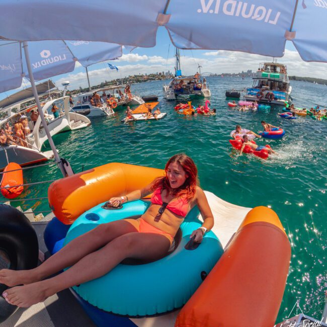 A woman in a pink bikini lounges on a large inflatable float at a lively boat party, surrounded by people in swimsuits relaxing on floats and swimming in the water under sunny skies.