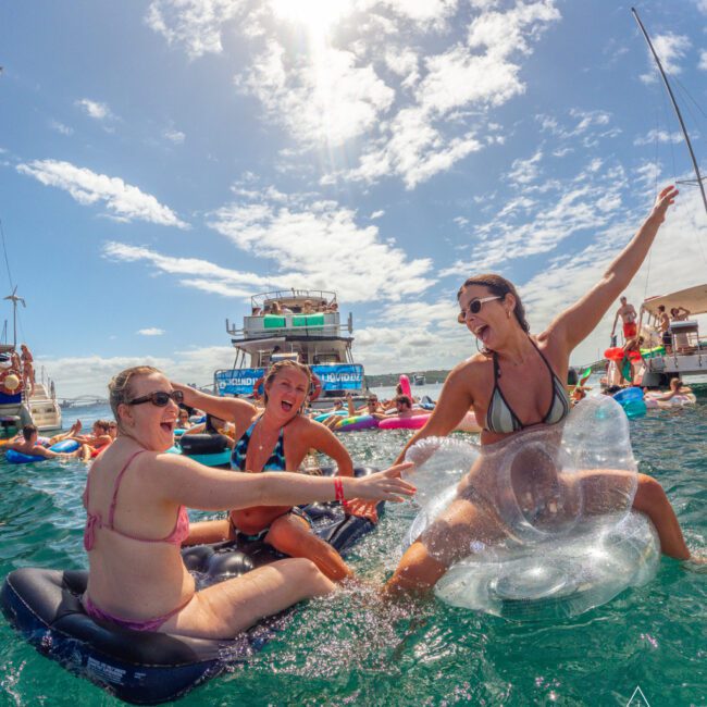 Three women laugh and play on inflatable floats in the ocean near boats under a bright, sunny sky. Other people are swimming and relaxing in the background. The scene is lively and fun.