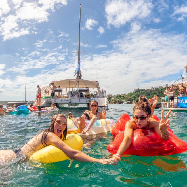 Three women in swimsuits relax on colorful pool floats, holding hands and smiling in the water near anchored boats on a sunny day. Other people are seen enjoying the water and the boat in the background.