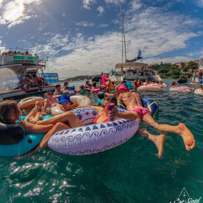 A group of people relax on colorful pool floats in the water near yachts and boats under a sunny sky, enjoying a lively social gathering. Some are chatting, while others lie back and soak up the sun.