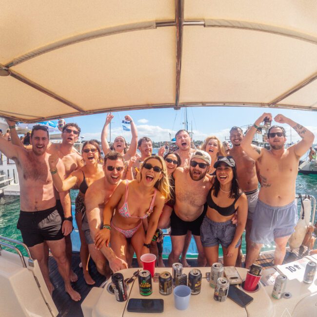 A group of excited young adults in swimwear pose and cheer on a boat under a canopy, surrounded by drinks and sunlight, with water and boats visible in the background.