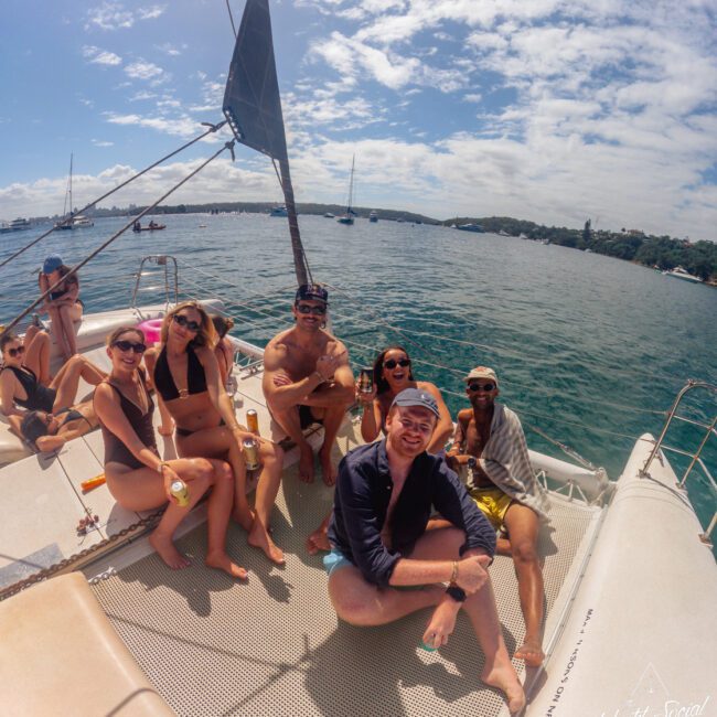 A group of seven people in swimwear relax and smile on the deck of a catamaran, with blue water, sailboats, and a partly cloudy sky visible in the background.
