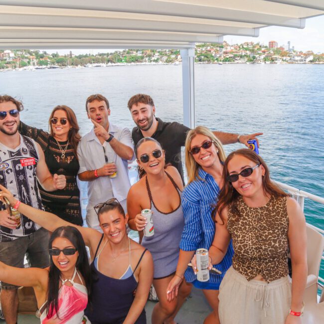 A group of nine smiling people pose together on a boat, holding drinks and wearing sunglasses, with water and a shoreline visible in the background under a sunny sky.