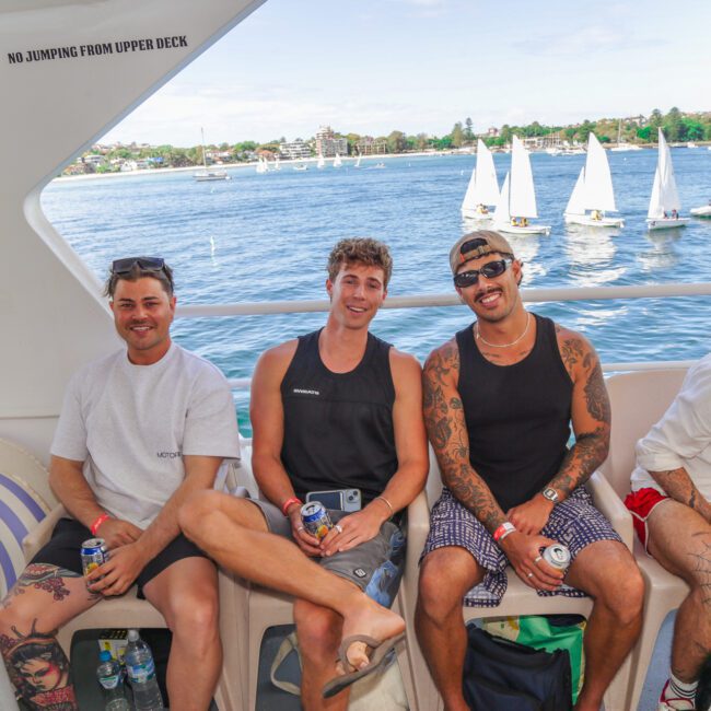 Three men sit and smile on a boat with drinks in hand, dressed in casual summer clothes. Behind them is a scenic view of water with sailboats and a distant shoreline under a clear sky.