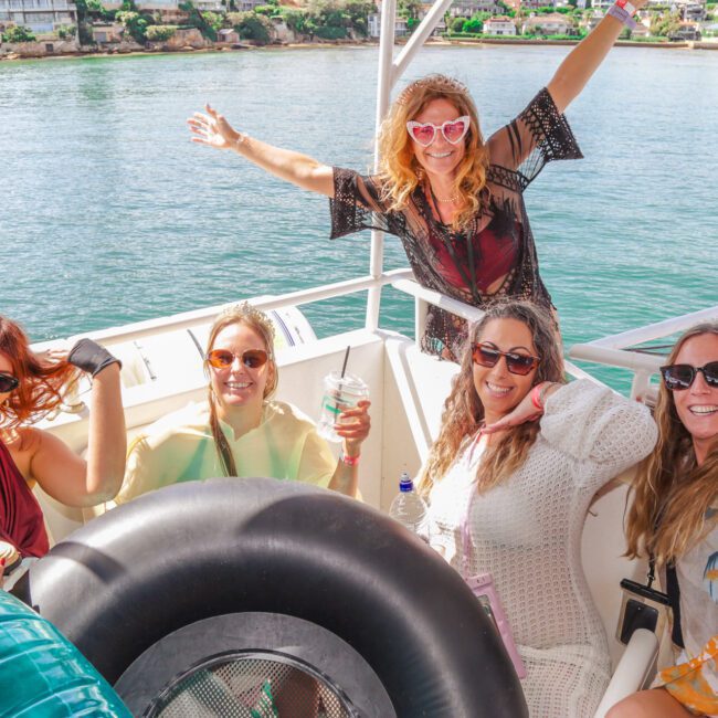 Five women smiling and posing on a boat, with water and houses onshore in the background. One woman stands with arms outstretched, and the others are seated, wearing sunglasses and summer outfits.