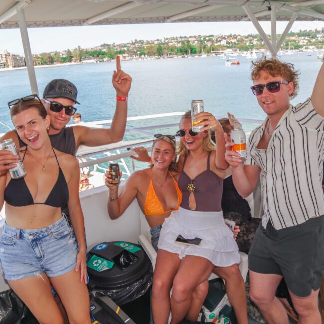 Five young adults in swimwear and sunglasses smile and raise drinks while posing on a boat. Water and a shoreline with houses are visible in the background under a sunny sky.