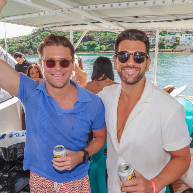 Two men wearing sunglasses and casual shirts smile at the camera while holding drinks on a sunny boat party, with people and a scenic waterfront visible in the background.