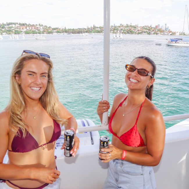 Two women in swimsuits smiling and holding drinks on a boat, with water, sailboats, and a cityscape in the background on a sunny day.