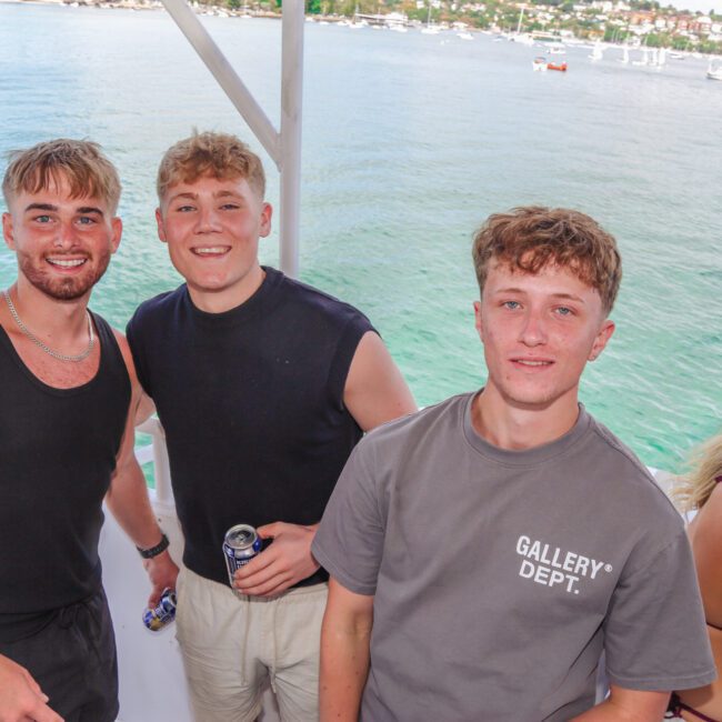 Three young men smiling at the camera on a boat, with water and sailboats in the background; one holds a drink can. A woman with blonde hair stands beside them, facing away. The scene is bright and summery.