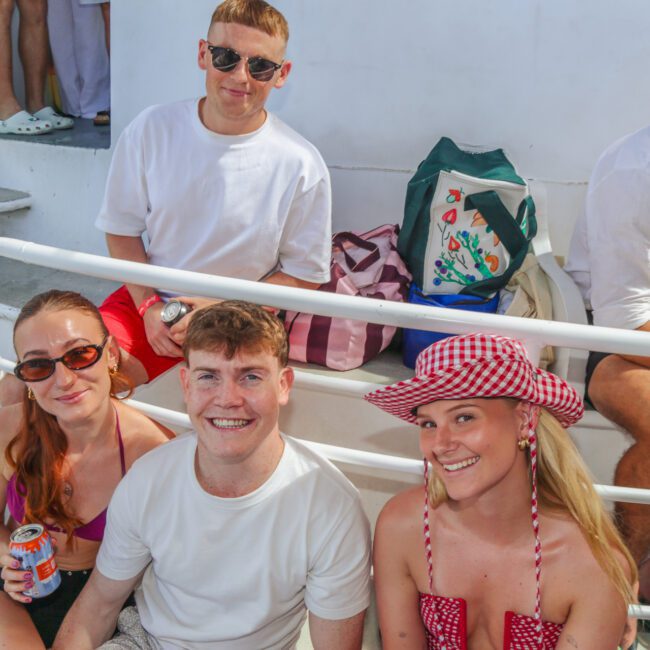 A group of four young adults, three sitting and one standing, smile at the camera on a boat. They wear casual summer clothes and sunglasses. A beach bag and drinks are visible beside them.