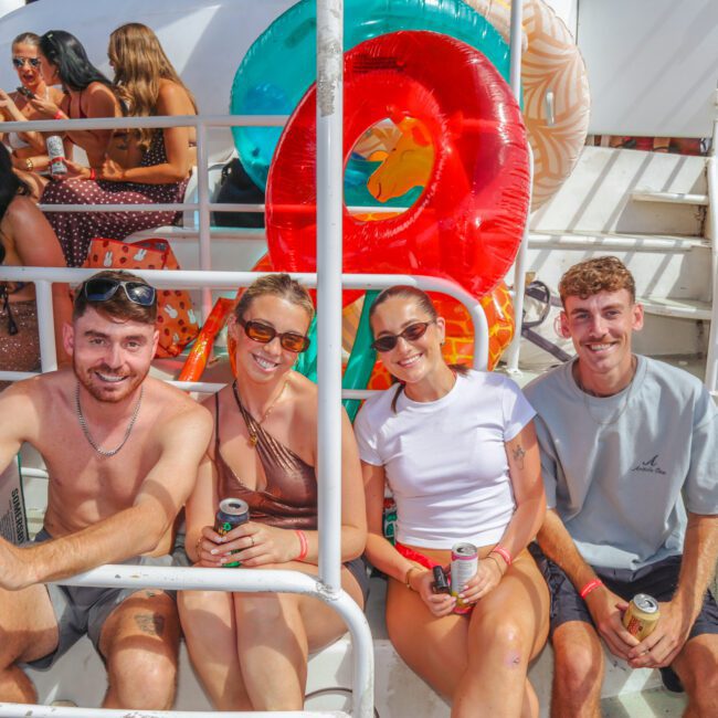Four smiling people sit on a boat’s steps holding drinks, with colorful pool floats behind them. Other people are in the background enjoying the sunny day on the boat. The ocean is visible to the right.