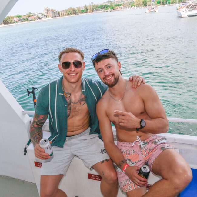Two men sit side by side on a boat, smiling at the camera. One wears sunglasses, a striped shirt, and shorts; the other is shirtless in pink shorts. Both hold drinks. The ocean and other boats are visible in the background.