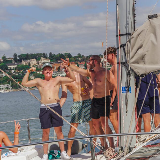 Four shirtless men stand and pose on a sailboat, smiling and gesturing playfully. The boat is on the water, with buildings and greenery visible in the background under a partly cloudy sky.