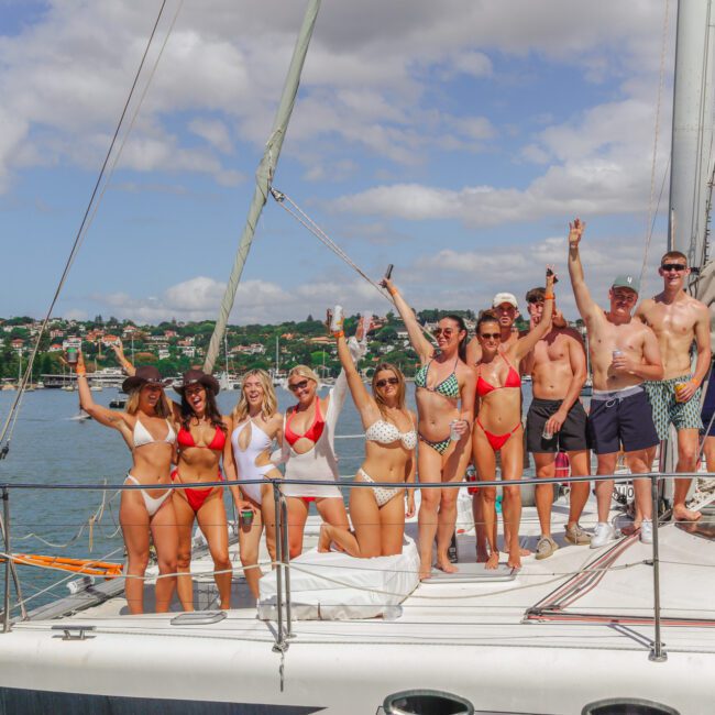 A group of people in swimsuits pose and smile on the deck of a sailboat anchored near the shore, raising their arms and enjoying a sunny day with water and a coastal town in the background.