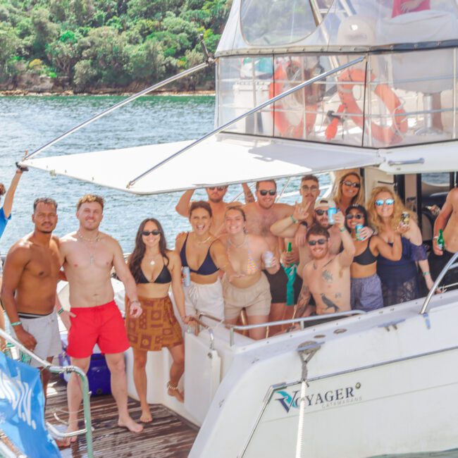 A group of people in swimwear smile and pose together on a docked white boat, surrounded by water and greenery, enjoying a sunny day. Some hold drinks, and a few people on a small boat wave in the background.