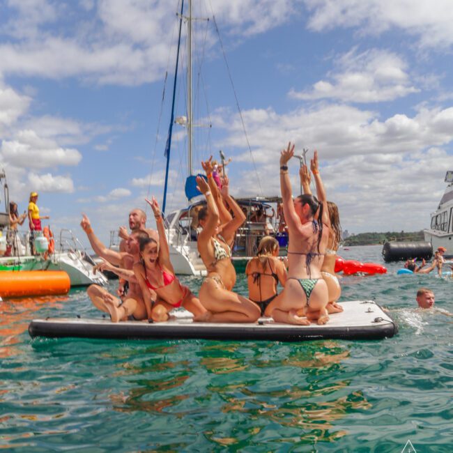 A group of people in swimsuits sit and cheer on a floating platform in clear blue water, with boats in the background under a partly cloudy sky. The scene appears lively and fun, suggesting a social gathering or party.