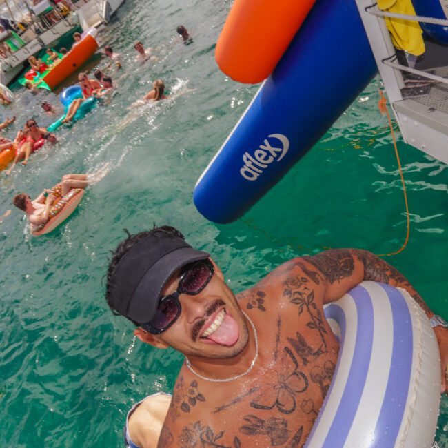 A smiling man with tattoos, sunglasses, and a cap floats on an inflatable ring in clear water, holding a drink. In the background, people on floats enjoy a lively party near boats. The scene is bright and festive.