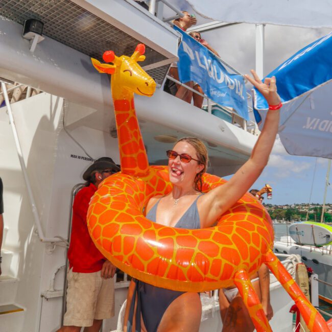 A smiling woman in a swimsuit stands on a boat, wearing large sunglasses and a giraffe-shaped pool float around her. She raises one arm in excitement, with people and a blue sky in the background.